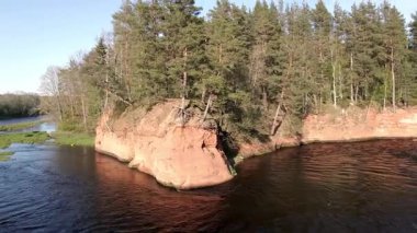 Uzun çam ağaçlarından oluşan sık bir ormanın tepesinde, kıvrımlı bir nehrin üzerinde kırmızı bir kumtaşı uçurumu yükselir. Sahne yumuşak, doğal ışıkla aydınlanır..