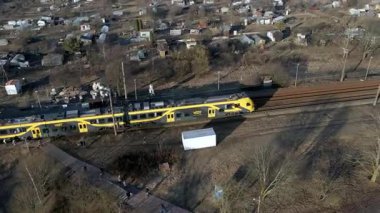 Modern bir sarı ve siyah tren bir demiryolu hattı boyunca seyahat eder. Nadir bitki örtüsü ve yapraksız ağaçlar, yakınlarda küçük bir yerleşim yeri bulunan kış aylarını akla getiriyor..