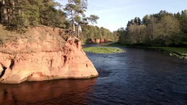 Ormanla çevrili kırmızı kumtaşı kayalıklarından bir nehir akar. Durgun su berrak gökyüzünü yansıtıyor, parlak gün ışığı sahneyi aydınlatıyor..