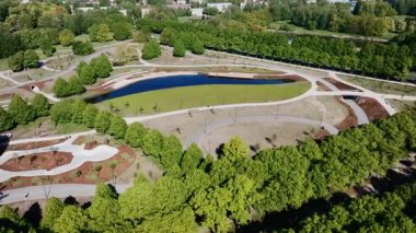 İyi korunan bir parkta oval bir gölet, yürüyüş yolları ve küçük bir köprü bulunur. Binalar ağaçların arasından kısmen görülebiliyor, bu da kentsel bir ortamı gösteriyor..