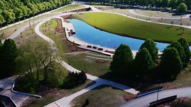 Bir park, yolu, genç ağaçları ve olgun ağaçlarla çevrili ahşap banklarla çevrili, gökyüzünü yansıtan kıvrımlı bir su özelliğine sahiptir..