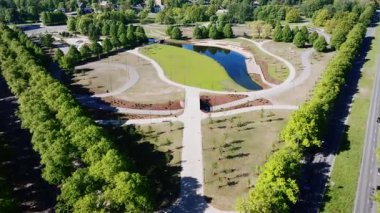 İyi korunan bir parkta oval şekilli bir gölet, yürüyüş yolları ve manikürlü çimenler, kentsel arkaplanda konut binaları görülebilir..