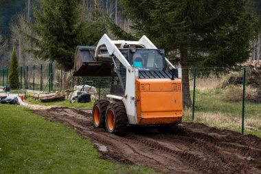 Küçük turuncu ve beyaz patinaj dümen yükleyicisi kirli bir yolda çalışır ve lastik izleri bırakır. Etrafı çitlerle çevrili, yemyeşil ağaçlar, kütükler ve çantalar..
