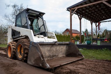 Çamurlu lastikleri olan bir patinaj yükleyicisi tahta bir pergola yakınındaki toprak bir patikada duruyor. İp ışıkları, dış mobilyalar, kırmızı bir ev ve çıplak ağaçlar..