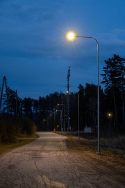Kırık bir yol mavi bir akşam gökyüzünün altında uzanır, sokak lambalarıyla kaplıdır. Uzun ağaçların yakınında bir telekomünikasyon kulesi duruyor..