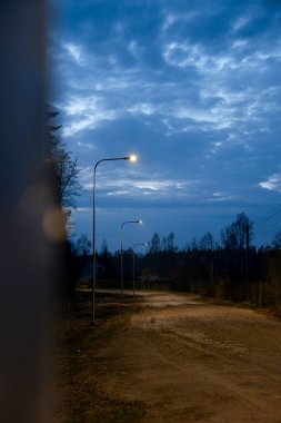 Bulutların dağıldığı loş bir gökyüzünün altında sokak lambalarıyla aydınlatılmış sessiz bir toprak yol. İnce ağaçlar ve uzak evler kırsal bir atmosfer yaratıyor..