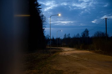 Sokak lambalarıyla kaplı toprak bir yol alacakaranlıkta sıcak bir parıltı yayar. İnce ağaçlar, çalılar, elektrik direkleri ve uzak ev siluetleri sahneyi tamamlıyor..