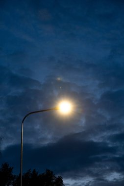 Bir sokak lambası parlak bir parıltı yayarak karanlık, bulutlu bir akşam gökyüzüne karşı bir halo etkisi yaratır. Ağaç yaprağı siluetleri kontrastı aşağıya ekler.