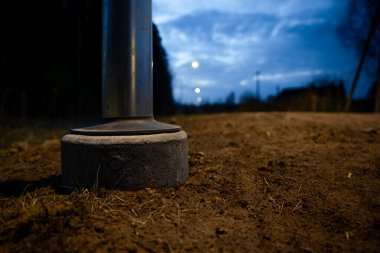 Beton temeli, soluk sokak lambaları, ağaçlar ve alacakaranlıkta kısmen bulutlu mavi bir gökyüzü olan toprak bir yolda metalik bir direğin tabanının alçak açısı..