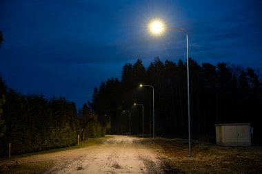 Alacakaranlıkta sokak lambalarıyla aydınlatılan toprak bir yol, bir tarafında uzun ağaçlar, diğer tarafında çit ve yanında da kullanılabilir bir alet kutusu olan bir çit..