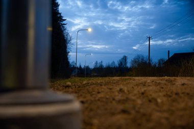 Derin mavi bir gökyüzünün altında sokak lambalarıyla aydınlatılan toprak bir yol. Elektrik hatları, elektrik direği, ağaç siluetleri ve görünürde bulanık metalik bir nesne var..