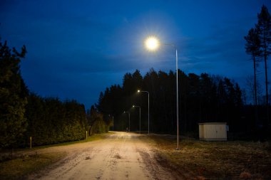 Sokak lambalarıyla aydınlatılmış, uzun ağaçlar ve çitlerle çevrili kırsal bir toprak yol. Sağda, koyu mavi bir gökyüzünün altında bir malzeme kutusu görülebilir..