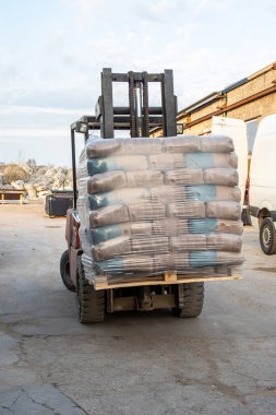 Forklift, beton zemin, tuğla bina ve park halindeki beyaz minibüslerin bulunduğu sanayi bölgesinde plastik poşet taşır..