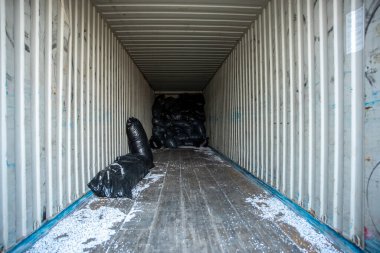 Beyaz duvarlı nakliye konteynırı, tırtıklı yapısı, ahşap zemin, en uçtaki siyah plastik torbalar, ve yere saçılmış beyaz parçacıklar..