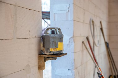 Koli bandına sarılı geçici bir cihaz, ahşap bir rafta, hafif tuğla bir duvarın karşısında duruyor. Etrafı aletlerle çevrili ve bitmemiş bir atölye ortamı var..