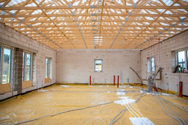 Açıkta duran ahşap çatı kirişleri, tamamlanmamış beton blok duvarları ve görülebilir elektrik ve su tesisatı kanalları olan bir binanın içi..