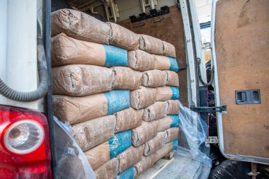 Kahverengi ve mavi çimento torbaları düzgünce istiflenmiş beyaz bir minibüsün arka kompartımanını aç. Tahta paneller ve kırmızı arka lamba görünür durumda. Plastik kaplama kargoyu kısmen kaplıyor..