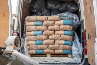 Tahta bir paletin üzerinde kahverengi ve mavi çimento torbalarıyla dolu bir minibüsün arkasında siyah plastik torbalar ve ön planda inşaat malzemeleri var..