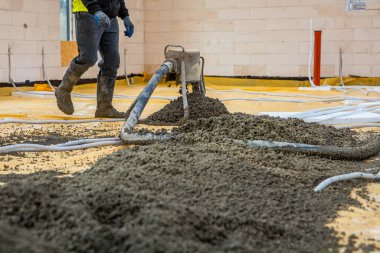 Botları ve eldivenleri olan bir işçi, yer altı ısıtma tesisatı için görünür boruları olan sarı tabakanın üzerine ıslak beton dökmek ve yaymak için ekipman kullanır..