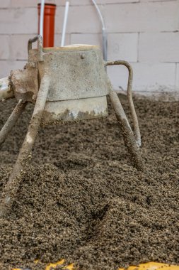 Çakıl taşlarının üzerinde beton karıştırıcısı olan bir inşaat alanı. Arka planda turuncu bir boru da dahil olmak üzere görünür boruları olan beyaz tuğlalı bir duvar..