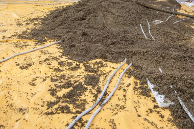 Bir inşaat alanı, kısmen çakıl veya beton karışımı ile kaplı sarı bir plastik levha gösteriyor, beyaz borular yüzeye saçılmış..