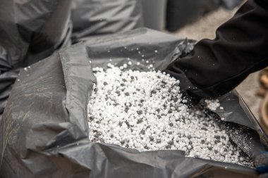 Eldivenli bir el, beyaz ve gri granüler malzeme içeren siyah bir plastik torbaya uzanır. Ek torbalar ve görünürde çakıl yüzeyi vardır..