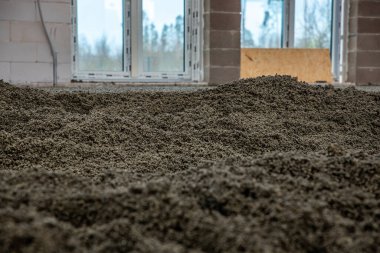 Zeminde çakıl taşları, beton blok duvarlar, geniş pencere aralıkları ve çerçeveye yaslanmış ahşap bir tahta olan inşaat alanı..