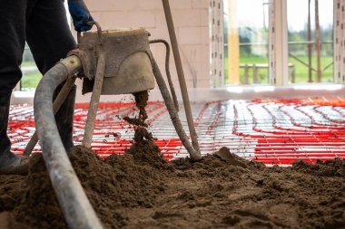 Bir işçi inşaat halindeki bir binanın içinde açık pencereli pompa kullanarak beton dökerken, yerde kırmızı ısıtma boruları düzenleniyor..