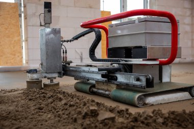 Beton zeminde kırmızı saplı, kum seviyesinde özel bir makine. Etrafındaki duvarlar tamamlanmamış, beton bloklar ve kontrplak açıkları var..