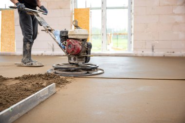 Bir işçi inşaat alanındaki beton zemini pürüzsüzleştirmek için bir güç malası kullanıyor. Yeraltı ısıtma boruları beton tabakanın altında görünür..