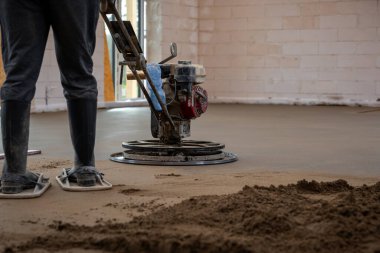 Bir işçi, beyaz tuğladan duvarları ve açık kapısı olan bir odada zeminin altındaki ısıtma borularının üzerindeki beton zemini pürüzsüz hale getirmek için bir güç malası kullanıyor..