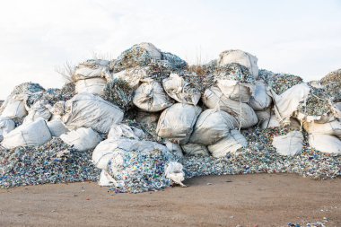 Plastik atıkla dolu büyük beyaz çuvallar, çıplak ve tozlu zemine saçılmış döküntüler, kirlilik ve atık birikimini vurguluyor..