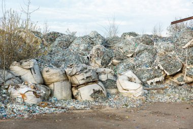 Parçalanmış büyük plastik atık yığınları ve yırtık çuvallarda atılmış şişeler. Etrafı yapraksız çalılar ve ağaçlarla çevrili..