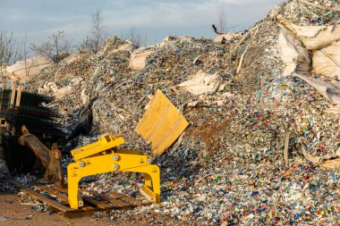 Çöp sahasında ya da çöp sahasında sarı bir endüstriyel makine bulunan şişe ve enkaz da dahil olmak üzere büyük bir plastik atık yığını. Uzakta seyrek ağaçlar.
