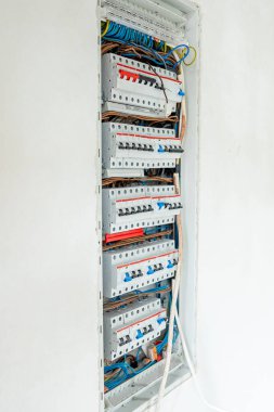 Beyaz bir duvardaki açık elektrik paneli düzenlenmiş devre kesicileri ve mavi ve sarı da dahil olmak üzere düzgün düzenlenmiş renk kodlu kabloları gösteriyor..