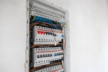 Beyaz bir duvara gömülü açık bir elektrik paneli düzgün düzenlenmiş devre kesiciler, konnektörler ve mavi, kahverengi ve sarı yeşil kabloları gösteriyor..