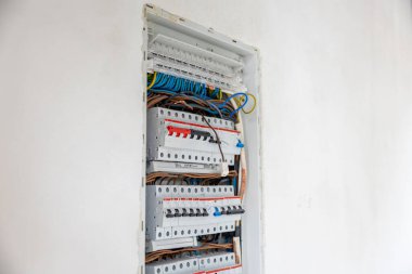 Beyaz bir duvara gömülü açık bir elektrik paneli devre kesicileri gösteriyor, mavi, kahverengi ve sarı yeşil kablolar düzenli ama tamamlanmamış bir durumda..