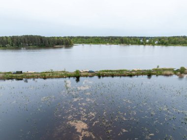 Su bitkileriyle yansıtıcı bir su yüzeyi, orman ve uzak yapılarla çevrili, seyrek bitki örtüsü olan dar bir kara şeridi ile bölünmüş..
