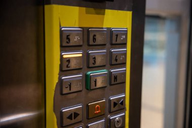 1 'den 7' ye kadar düğmeli metalik asansör kontrol paneli, kabartma yazılar, yeşil çizgili 1. kat düğmesi ve aşınmış sarı kenar çizgisi..