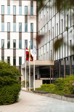 Alternatif beyaz ve koyu panelleri olan modern ofis binası, üç bayrak direği, asfalt yol ve B girişine yakın ön planda bitkiler..