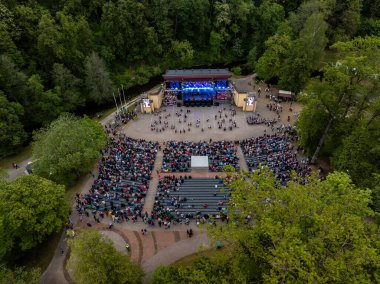 Uzun ağaçlarla çevrili bir açık hava amfi tiyatrosunun havadan görünüşü. Mavi ışıklı bir sahne, büyük ekranlar ve bir seyirci topluluğu..