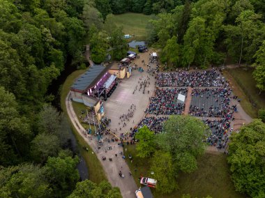 Bir açık hava amfi tiyatrosu, girişin yakınında ağaçlar, patikalar, akarsu ve görünür araçlarla çevrili oturma izleyicileri ile bir performans sunar..