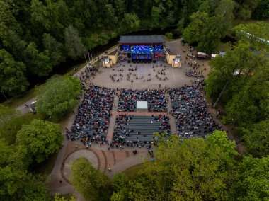 Ağaçlarla çevrili bir açık hava amfi tiyatrosunun hava görüntüsü. Mavi ışıklandırmalı, oturaklı ve belirgin dairesel yolları olan bir sahne..