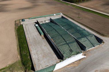 Yeşil brandaları ve beyaz çantaları olan, pullu tarlalarla çevrili bir toprak depolama tesisi ve üst çerçevede asfaltlı bir yol..