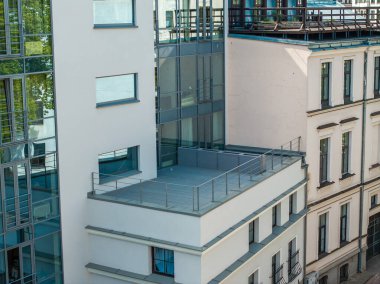 Geniş camlı pencereleri ve terası olan modern bir bina geleneksel krem rengi bir yapının yanında duruyor..