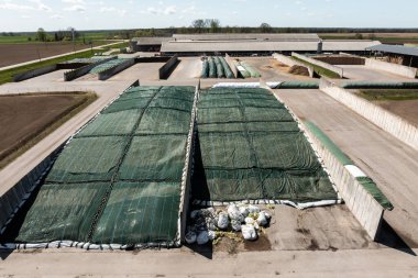 Yeşil branda ve beyaz kum torbalarıyla kaplı, parçalı bulutlu bir gökyüzünün altında açık tarım arazileriyle çevrili büyük tarımsal tesisler..