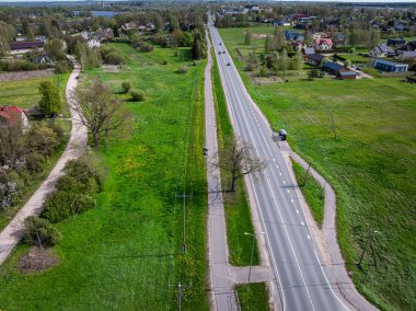 Açık araziler, dağınık evler, ağaçlar, elektrik direkleri ve paralel yaya yolu ile çevrili iki şeritli düz bir yolun hava görüntüsü..