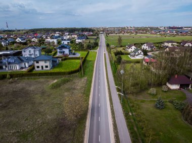 Evlerle, açık arazilerle ve seyrek ağaçlarla çevrili düz asfalt bir yol bulutlu bir gökyüzünün altında görünen bir kuleye sahip binalardan oluşan uzak bir kümeye çıkıyor..
