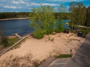 Slaytlı ve salıncaklı kumlu bir oyun alanı durgun bir su kütlesinin yanında oturur, tahta kaldırımlar, sazlıklar ve taze yeşil yapraklı ağaçlarla çevrili..