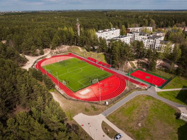 Kırmızı koşu pisti, yeşil futbol sahası, basketbol sahası, sık orman ve yakınlardaki yerleşim alanlarının yer aldığı bir spor kompleksinin havadan görünüşü.